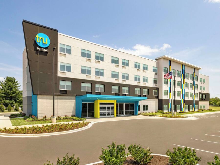 A modern hotel building with a prominent 'tru by Hilton' logo, a covered entrance, and a parking lot.