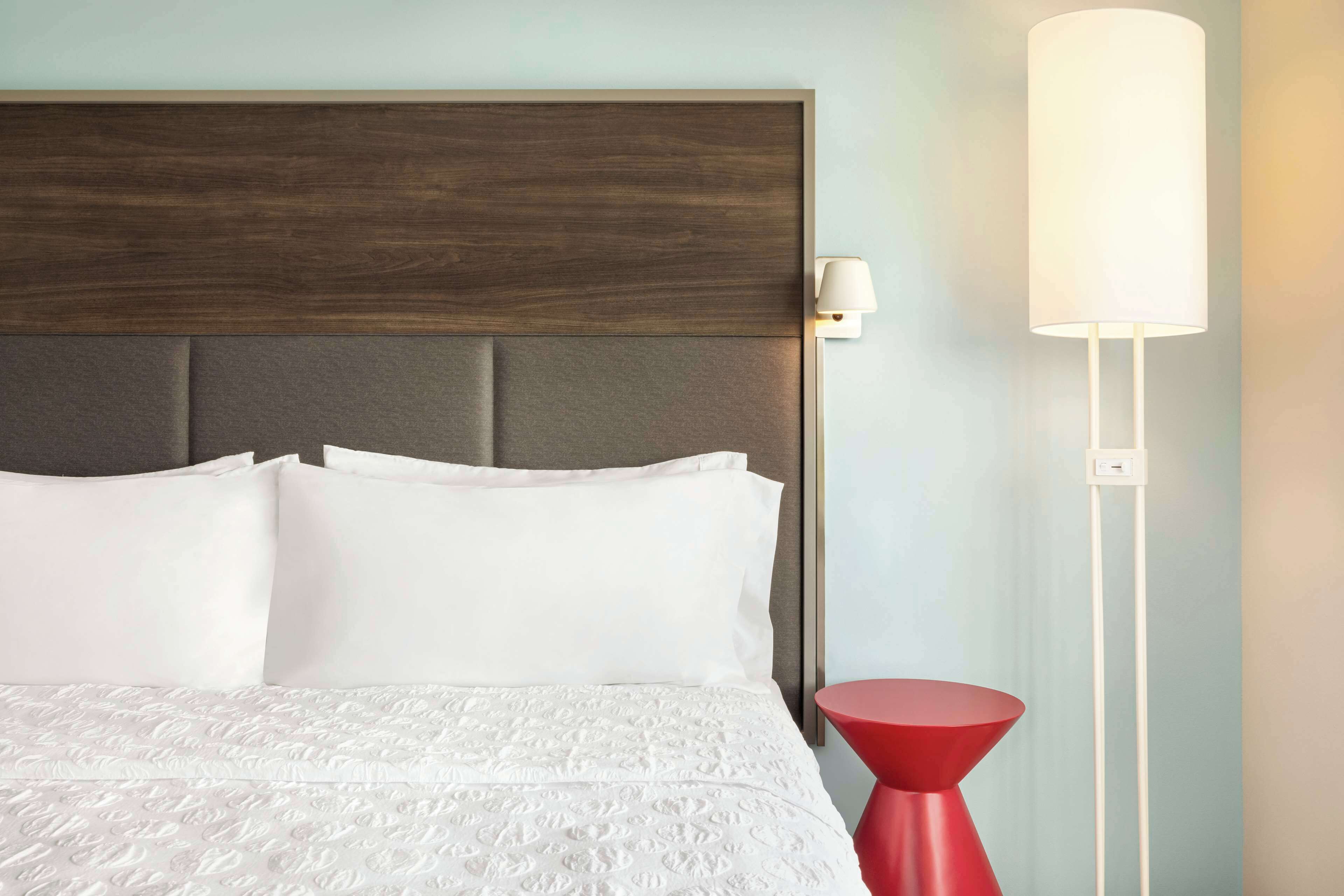 A neatly made bed with white linens, a wooden headboard, and a red side table with a lamp.
