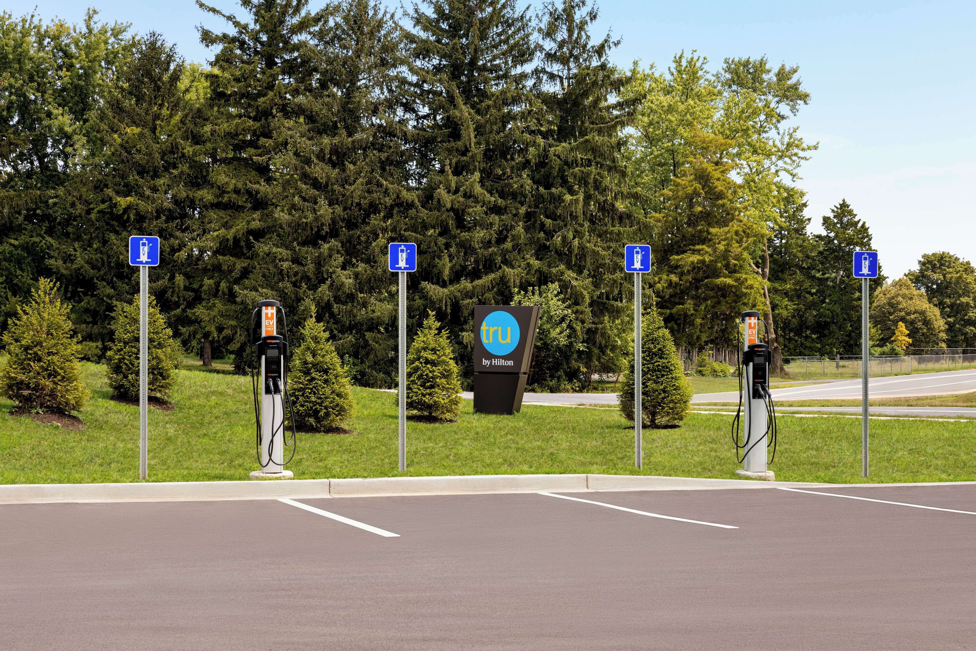 A parking area with electric vehicle charging stations, a sign for Hilton by Hilton, and a large tree in the background.