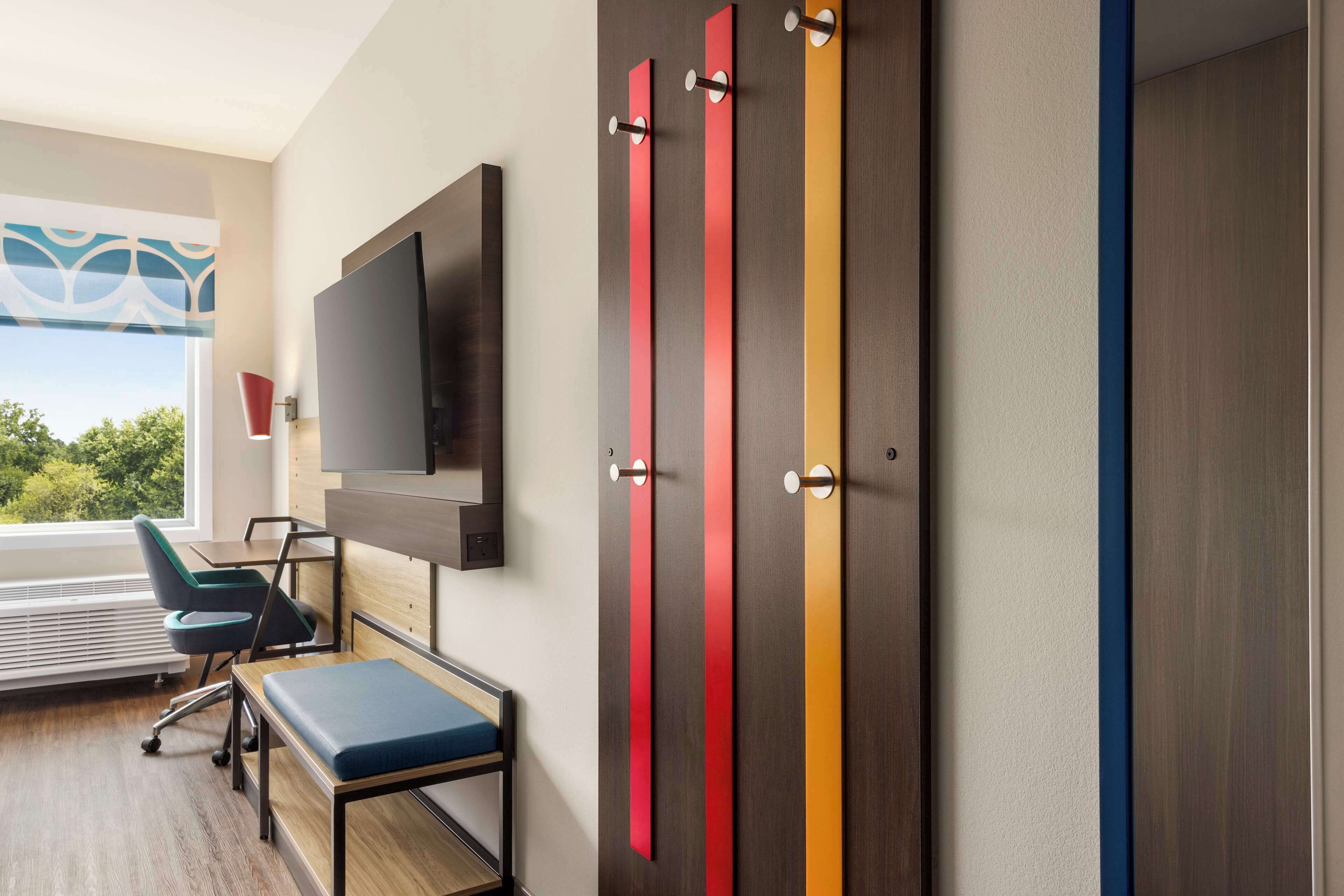 Habitación de hotel moderna con un televisor de pantalla plana grande montado en la pared, un escritorio con una silla, un banco y una ventana con vistas a zonas verdes.