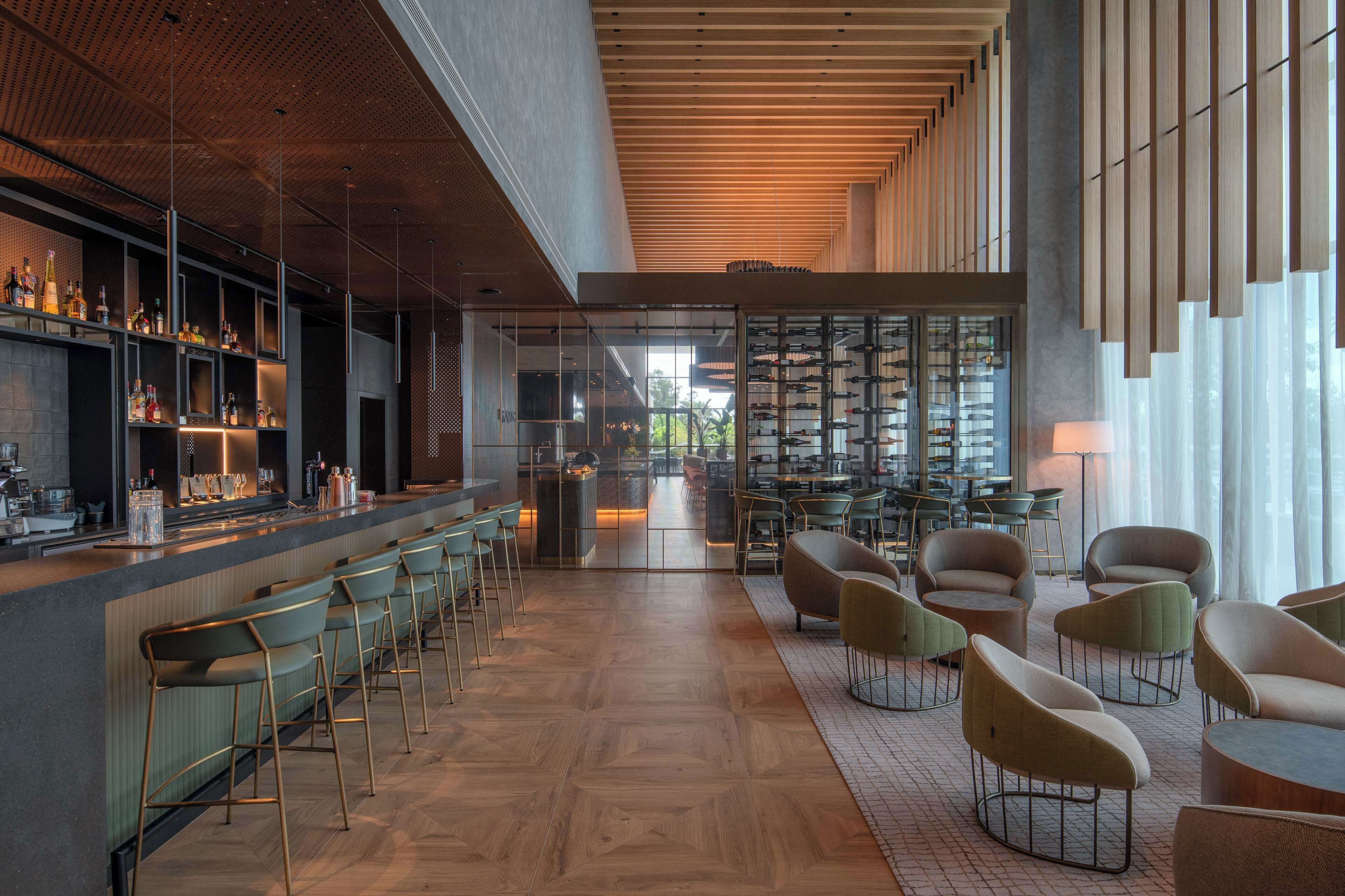 A modern bar area with a long counter, bar stools, and a variety of bottles. There are multiple seating areas with chairs and tables, and a large window providing natural light.