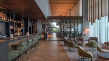 Un bar moderno con una barra larga, taburetes y una variedad de botellas. Hay varias zonas de estar con sillas y mesas, y un gran ventanal que proporciona luz natural.