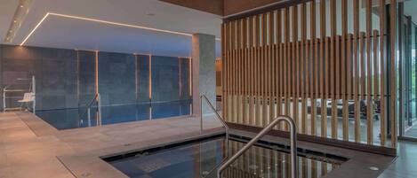 Un espace piscine intérieur moderne, doté d’une paroi en verre, de rampes métalliques et offrant une vue sur l’extérieur.