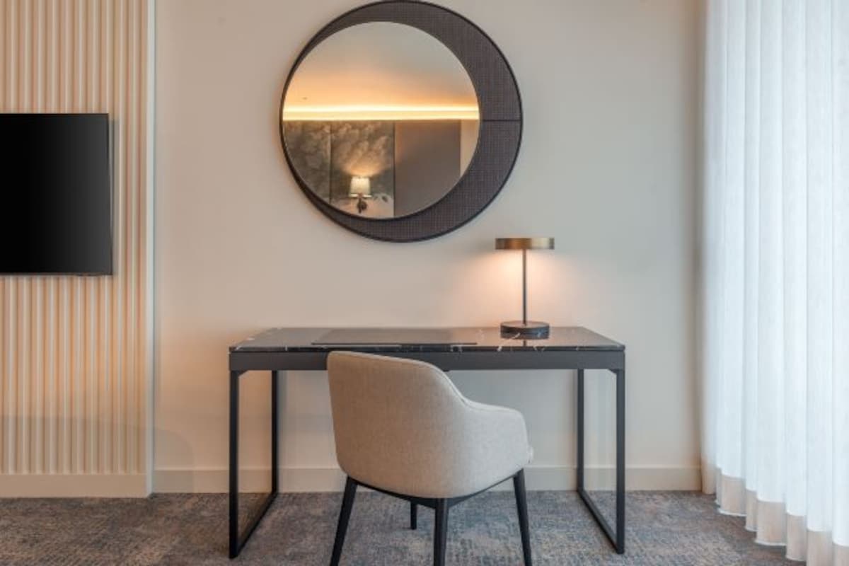 A modern room with a round mirror, a desk, a chair, a TV, and a lamp.