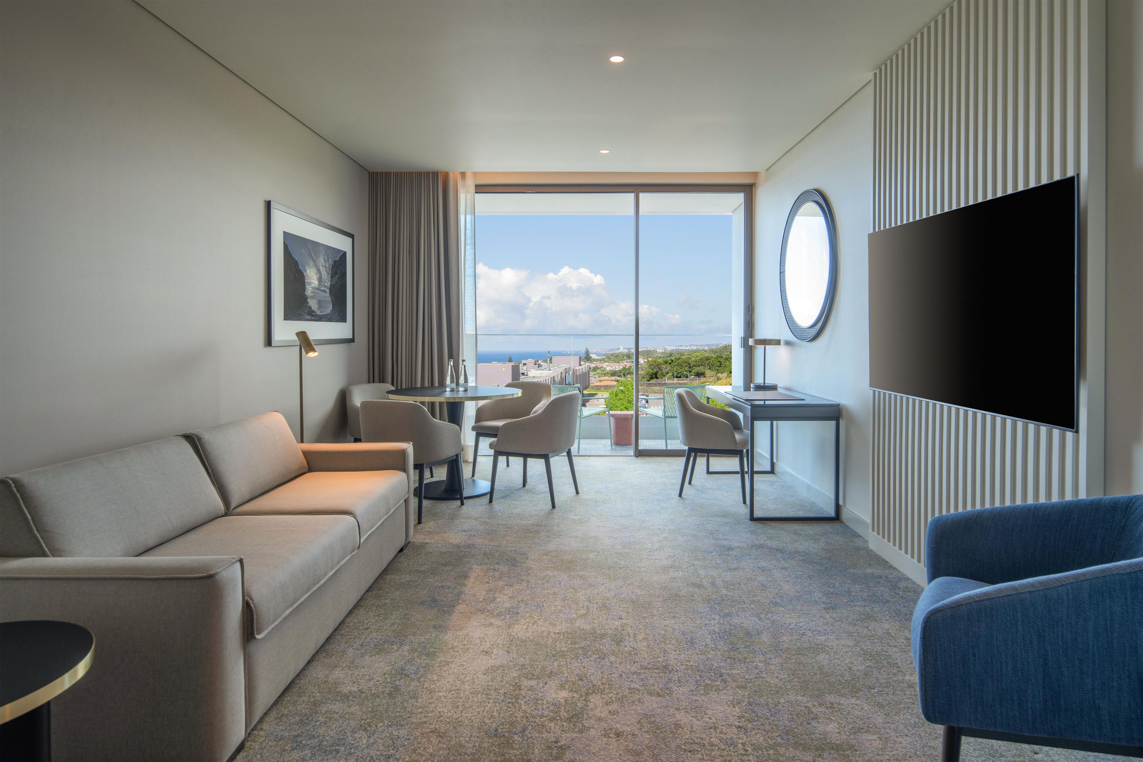 A hotel room with a sofa, a desk with chairs, a television, and a large window offering a view of the city.