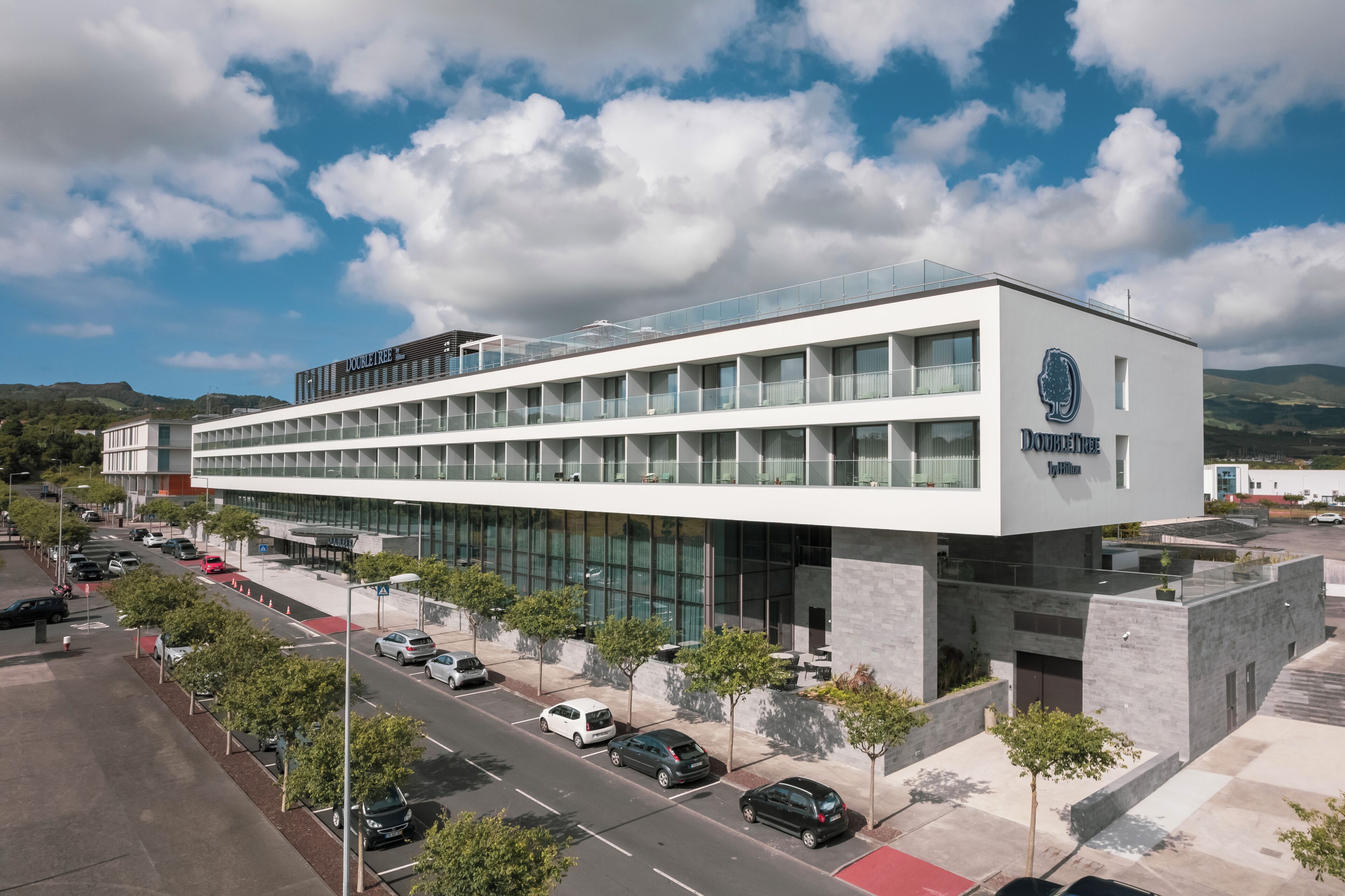 A modern building with a glass facade, multiple floors, and a parking lot with cars.