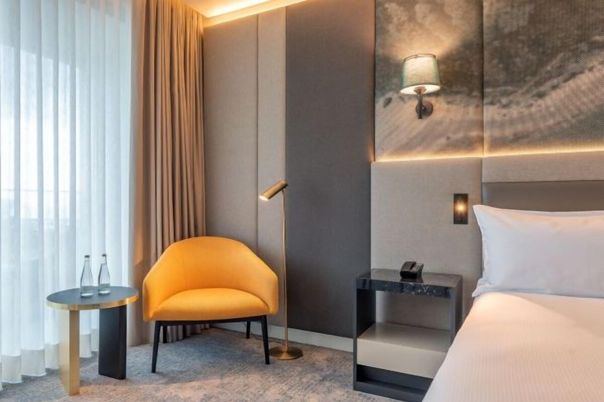 Una habitación de hotel con cama, una silla, una mesita, una mesita de noche y una lámpara.