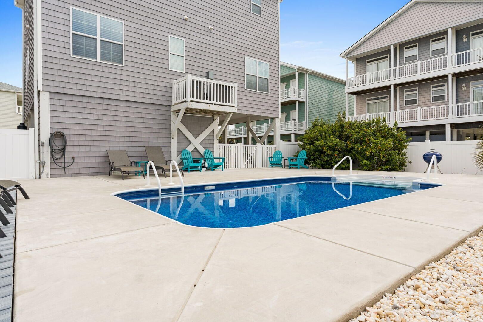 A swimming pool with a white deck, surrounded by chairs and a small deck area.
