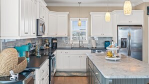 A modern kitchen with white cabinets, a stainless steel refrigerator, and pendant lights. - Rainbow Skies- 5 bedroom, 4 bath w/private pool, OIB resort & steps to the beach (Ocean Isle Beach)