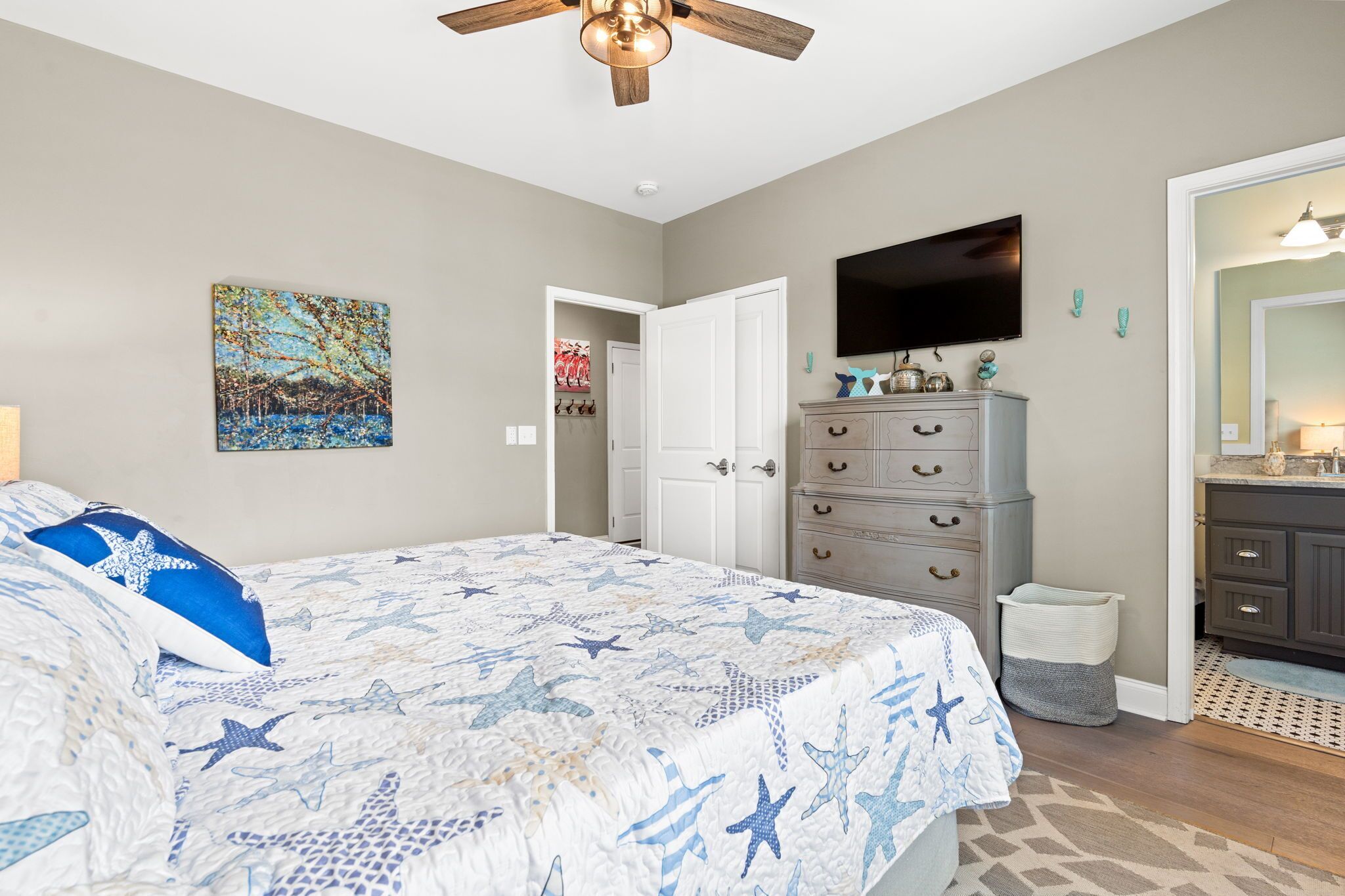 A bedroom with a bed, a ceiling fan, a TV, a dresser, and a bathroom visible through an open door.