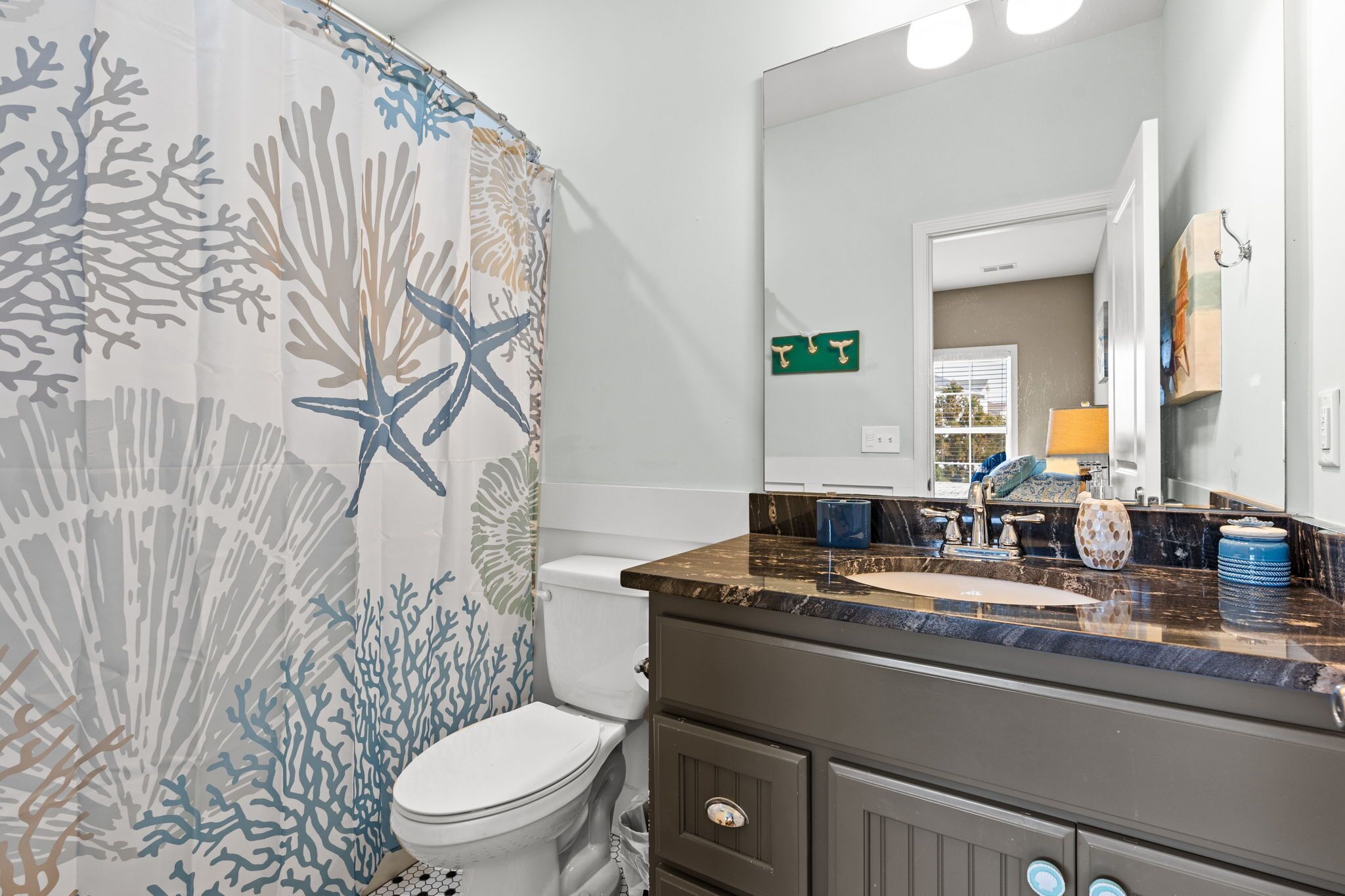 A bathroom with a shower curtain featuring a coral and starfish pattern, a white toilet, a vanity with a marble countertop, and a mirror above the sink.
