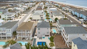 A coastal residential area with multiple two-story houses, a swimming pool, and a beach in the background. - Rainbow Skies- 5 bedroom, 4 bath w/private pool, OIB resort & steps to the beach (Ocean Isle Beach)