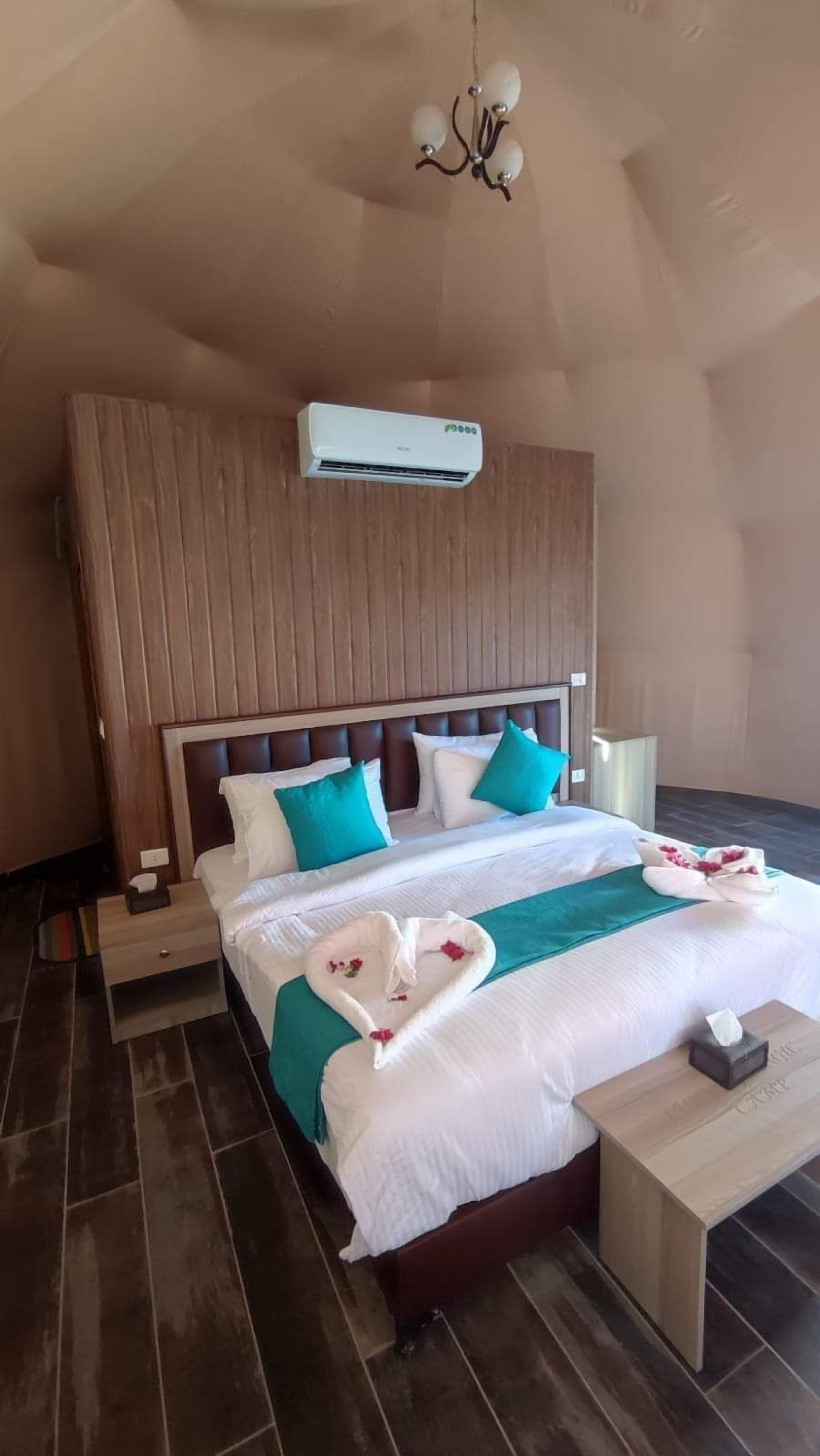 Una habitación de hotel con cabecera de madera, una cama con ropa de cama blanca y aguamarina, una toalla con forma de corazón y una mesita de noche pequeña.