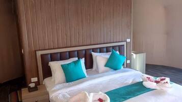 Una habitación de hotel con cabecera de madera, una cama con ropa de cama blanca y aguamarina, una toalla con forma de corazón y una mesita de noche pequeña.