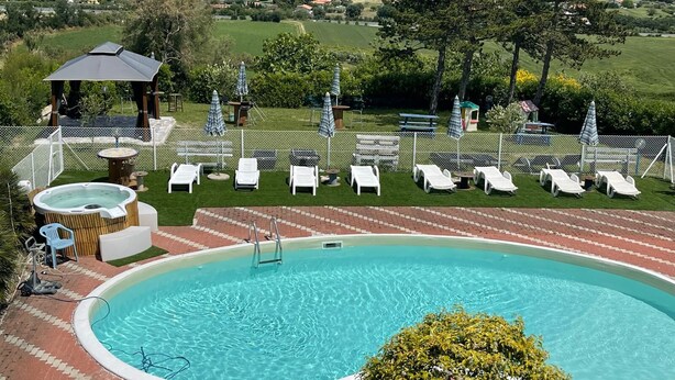 A swimming pool with lounge chairs and a hot tub, surrounded by a fence and greenery, overlooking a coastal town. - B&B Villa Alma (San Costanzo)