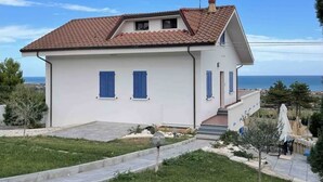 A single-story house with a tiled roof, white walls, and blue shutters. There is a small garden area with grass and a few trees. - B&B Villa Alma (San Costanzo)