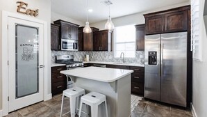 A modern kitchen with dark wood cabinets, stainless steel appliances, a white island with stools, and a pendant light. - 3 Bed Single Floor Sleeps w/pool (Washington)
