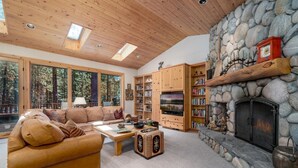 A cozy living room with a stone fireplace, wooden bookshelves, and a large window overlooking a forest.
