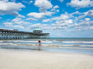 Une plage avec une jetée qui s’avance dans l’océan, une personne debout dans une eau peu profonde et un ciel dégagé avec des nuages.