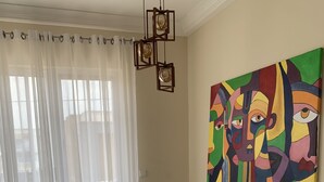 Een eethoek met een glazen tafel en witte stoelen, een kleurrijk abstract schilderij aan de muur en een uitzicht door dunne gordijnen.