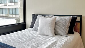 Un letto rifatto con una coperta bianca e una coperta in blu scuro, una finestra con vista su un edificio e una parete spoglia.