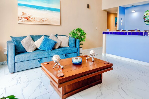 Un salon avec un canapé bleu, une table basse agrémentée d’objets décoratifs, une plante et un tableau représentant une scène de plage accroché au mur.