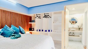 Ein modernes Schlafzimmer mit einem großen Bett, blauen Kissen, einem Holz-Kopfschild, einem Kleiderschrank und einem Badezimmer mit Spiegel und Waschbecken.