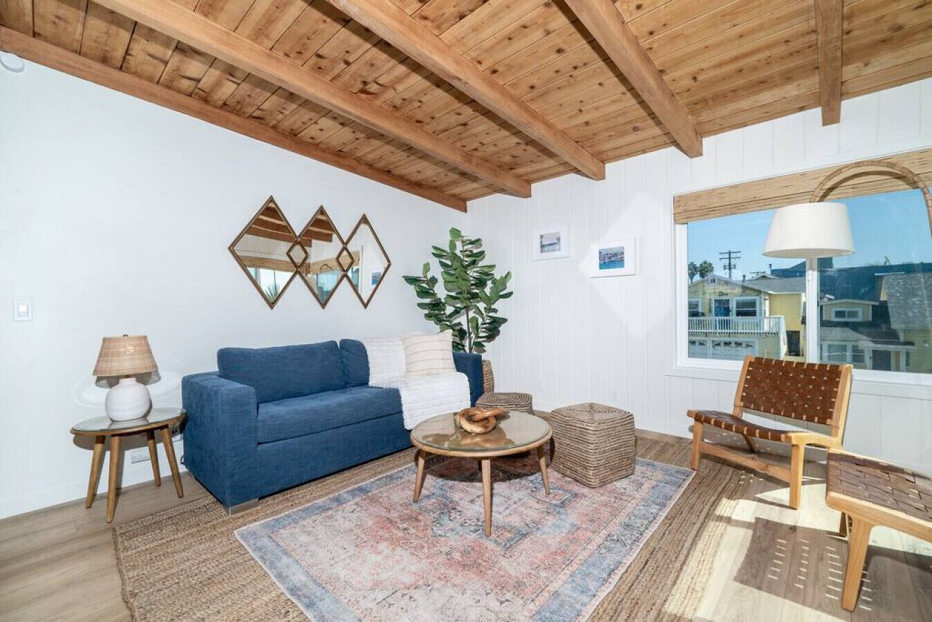 A living room with a blue sofa, a round coffee table, a wicker ottoman, a wooden bench, and a large window with a view of buildings.
