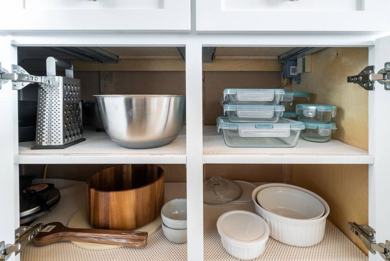 A kitchen cabinet with a grater, a large metal mixing bowl, stackable storage containers, a wooden bowl, and ceramic dishes.