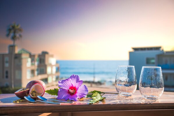 A table with two glasses, a passion fruit, and a hibiscus flower on a wooden surface with a view of the sea and buildings in the background.
