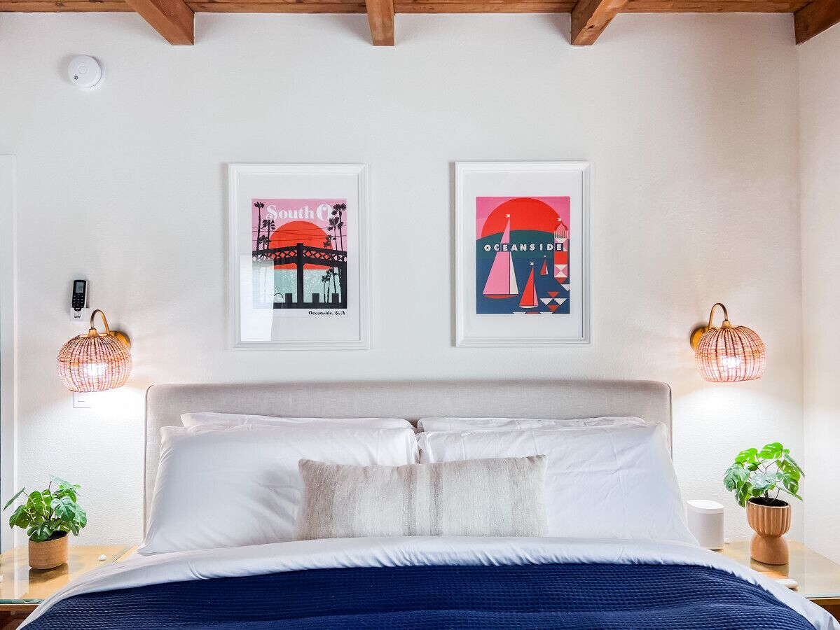 A bedroom with a bed, two wall-mounted artworks, bedside lamps, and a potted plant.