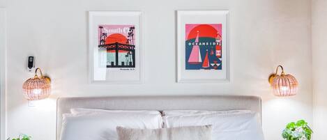 A bedroom with a bed, two wall-mounted artworks, bedside lamps, and a potted plant.
