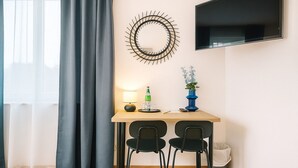 Habitación de hotel con un escritorio, dos sillas, un televisor, un reloj de pared, una lámpara, una botella y un florero con flores.