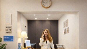 Un mostrador de recepción con un reloj, un cartel que indica los servicios sanitarios, y cuadros en la pared.