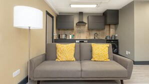 A modern apartment with a grey sofa, two yellow patterned cushions, a floor lamp, a microwave, a washing machine, and dark cabinets. - Ideally located cosy flat (London)