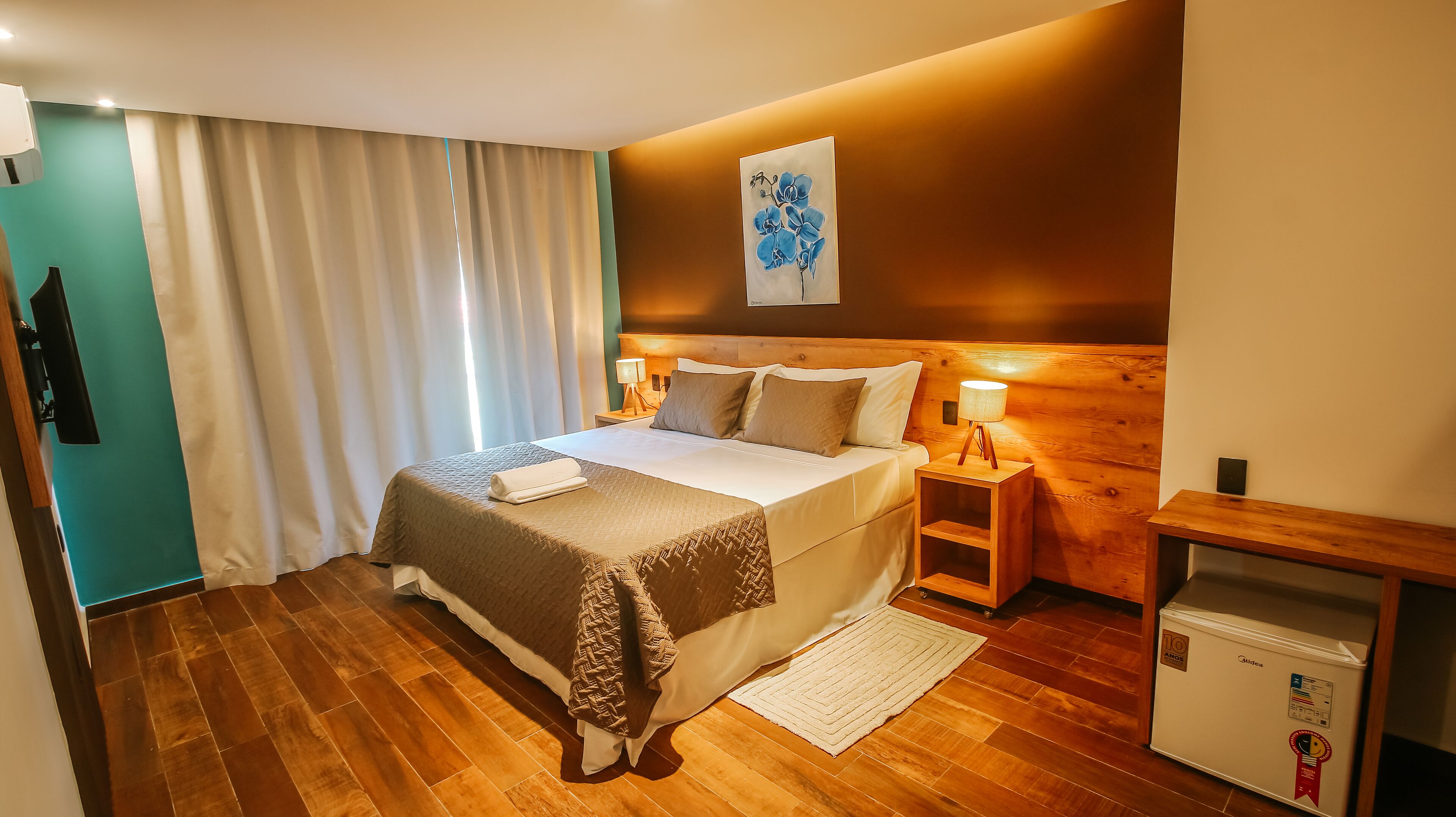 A hotel room with a wooden floor, a bed with a beige and brown bedspread, a nightstand with a lamp, and a small refrigerator.