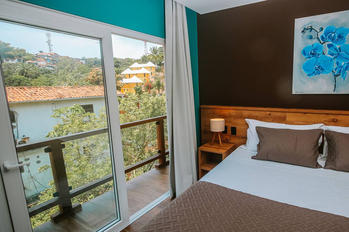 A hotel room with a bed, a nightstand, a painting, and a view of the outside.