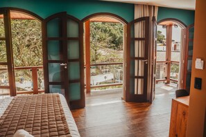 A bedroom with a large bed, wooden floor, and a balcony with a view of trees and buildings. - Pousada Flores da Praça (Cairu)