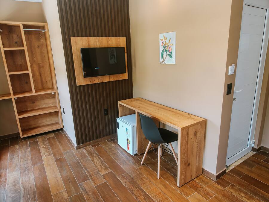 A room with a wooden desk, a TV mounted on a wooden panel, a white refrigerator, and a wooden shelving unit.