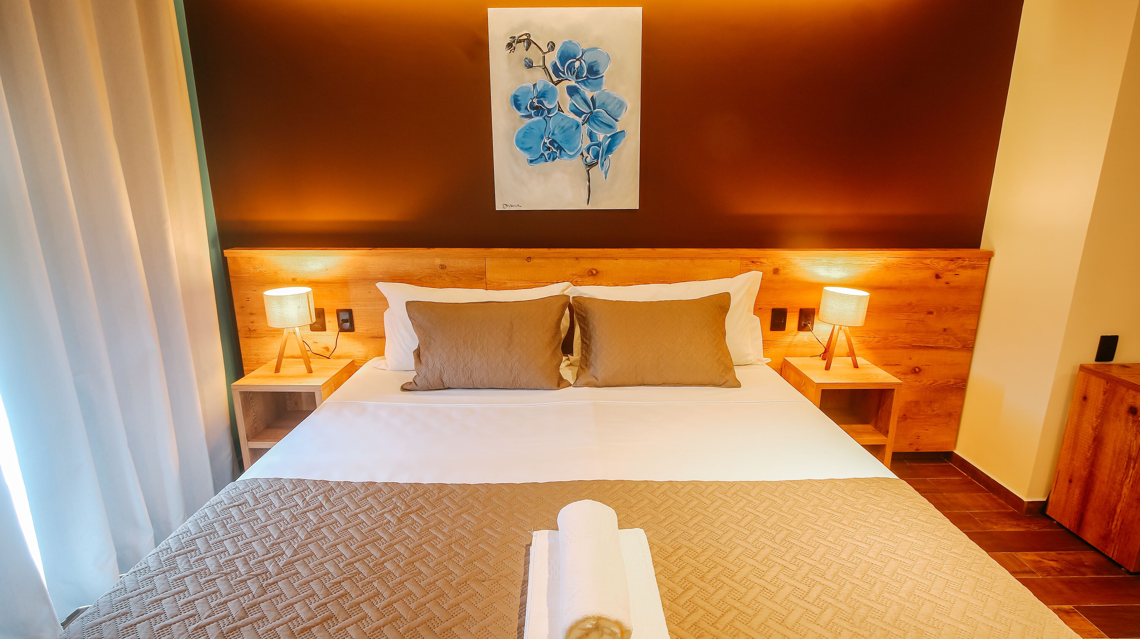 Une chambre d’hôtel avec une tête de lit en bois, deux tables de chevet avec des lampes, un lit avec du linge de lit blanc et un tableau représentant des fleurs bleues accroché au mur.