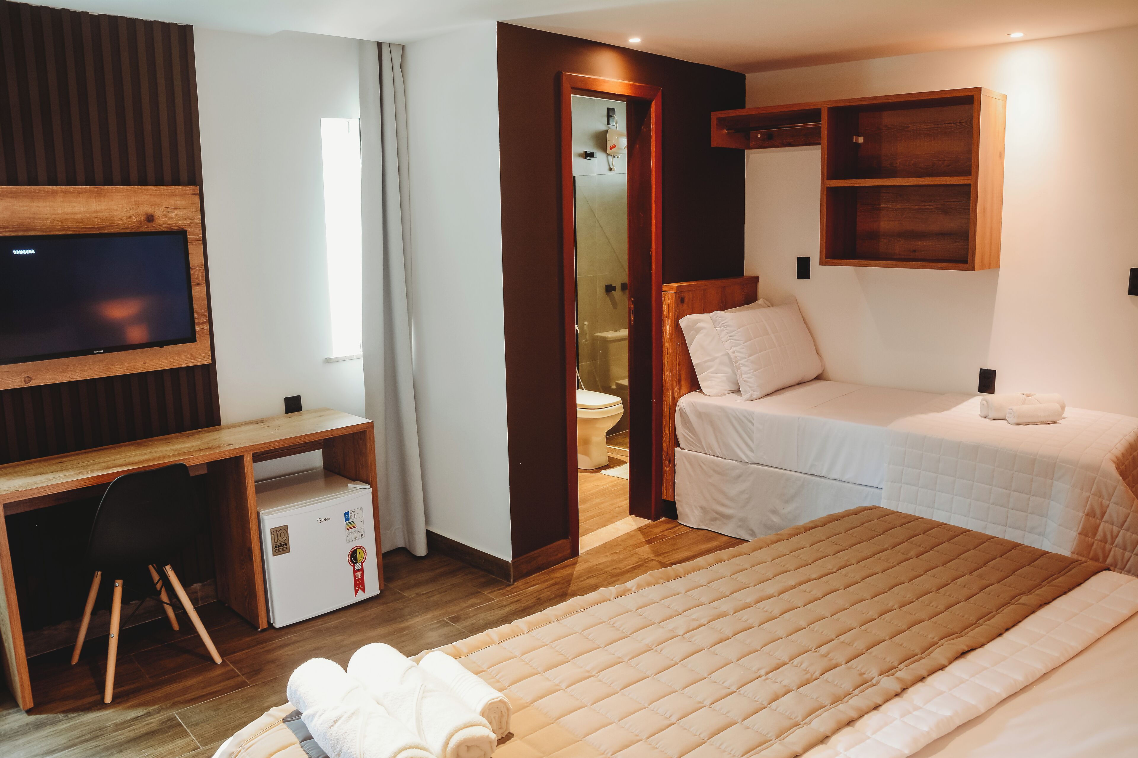 A hotel room with a bed, a desk with a TV, a small fridge, and a wooden shelf.