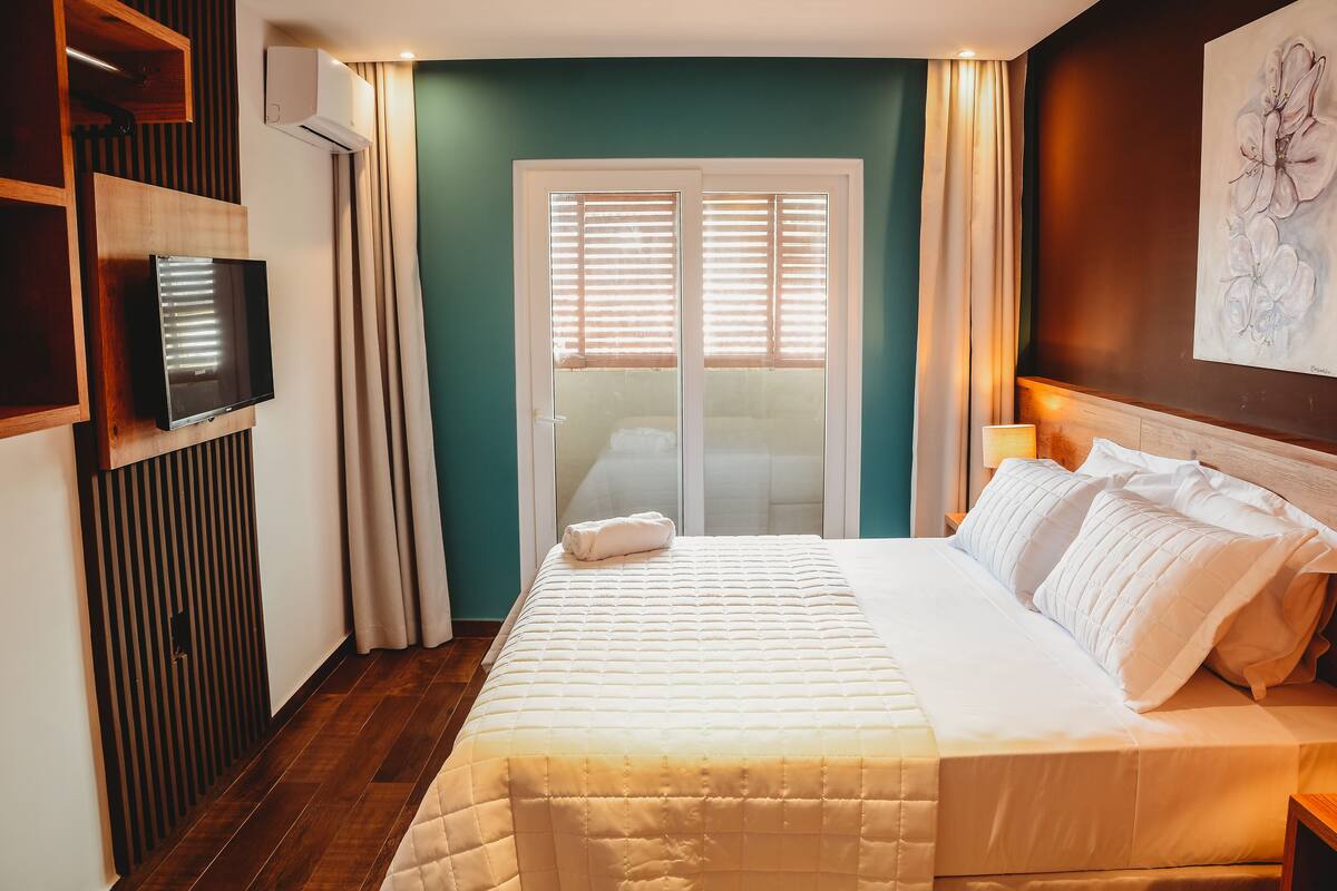 A hotel room with a bed, a TV mounted on the wall, a window with blinds, and a painting on the wall.