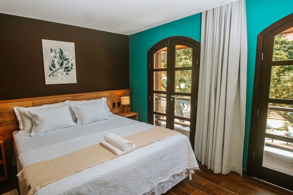 A neatly made bed with white linens and a beige throw blanket, a wooden headboard, a bedside table with a lamp, and a window with curtains.