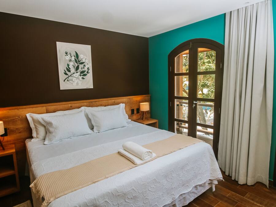 A neatly made bed with white linens and a beige throw blanket, a wooden headboard, a bedside table with a lamp, and a window with curtains.