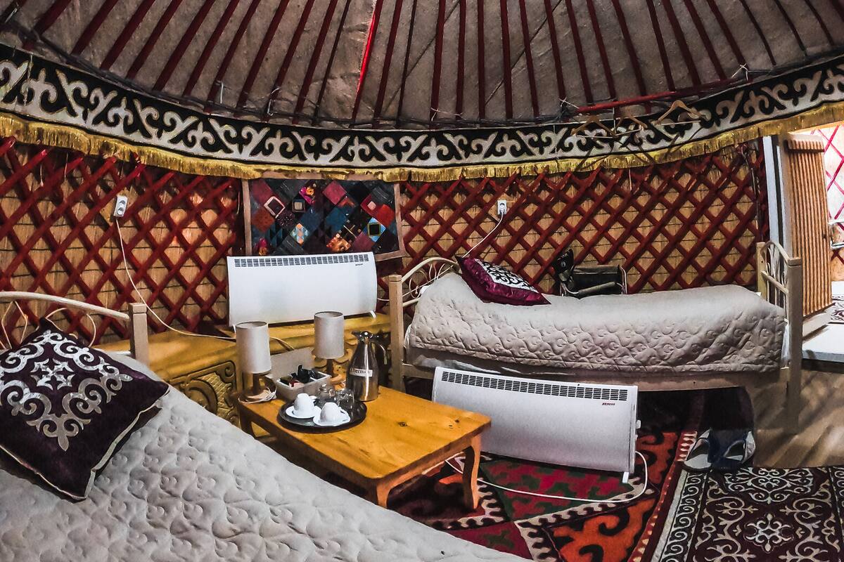 Interior de una yurta con techo abovedado, una cama, una mesa con hervidor y un pequeño calentador.