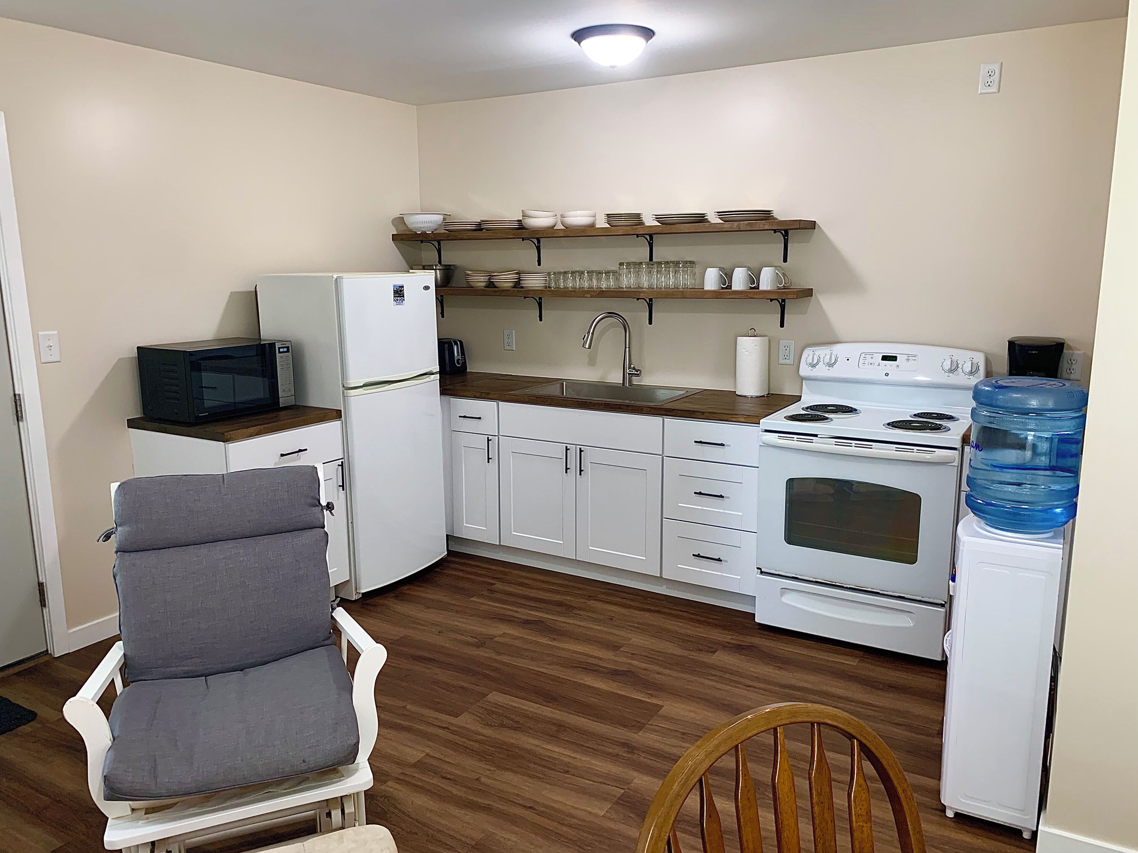 A compact kitchen with white appliances, a wooden chair, and a water dispenser.