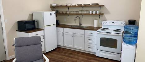 A compact kitchen with white appliances, a wooden chair, and a water dispenser.