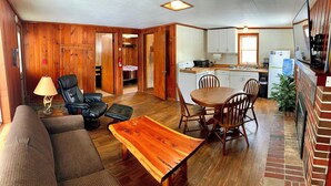 Ein Wohnzimmer mit einer Couch, einem Holztisch, einem schwarzen Ledersessel und einem Esstisch mit Stühlen. Es gibt eine Küchenzeile mit Schränken und einem Kühlschrank.