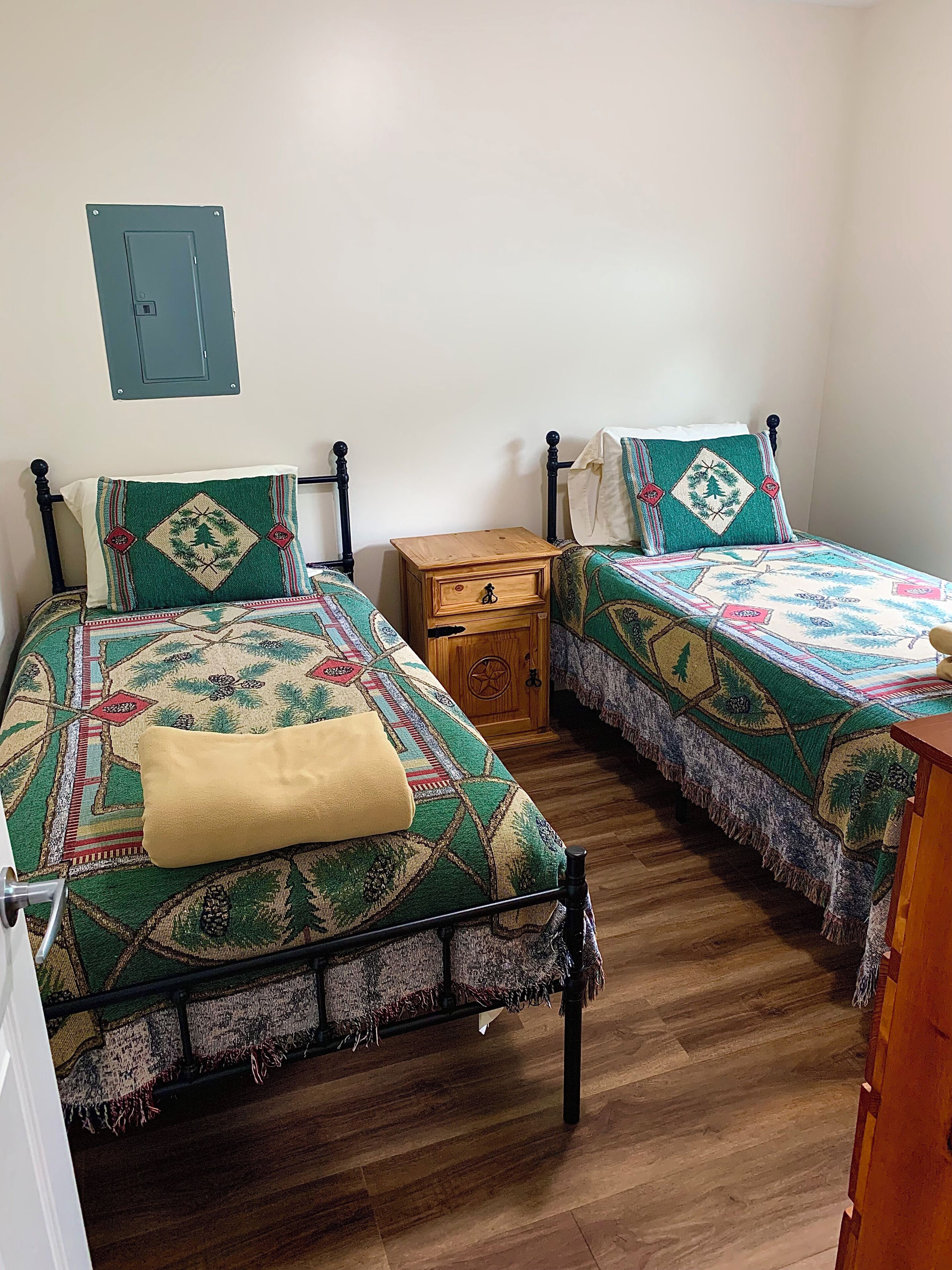 A room with two beds, each with a patterned bedspread and pillows, a wooden nightstand, and a wall-mounted electrical box.