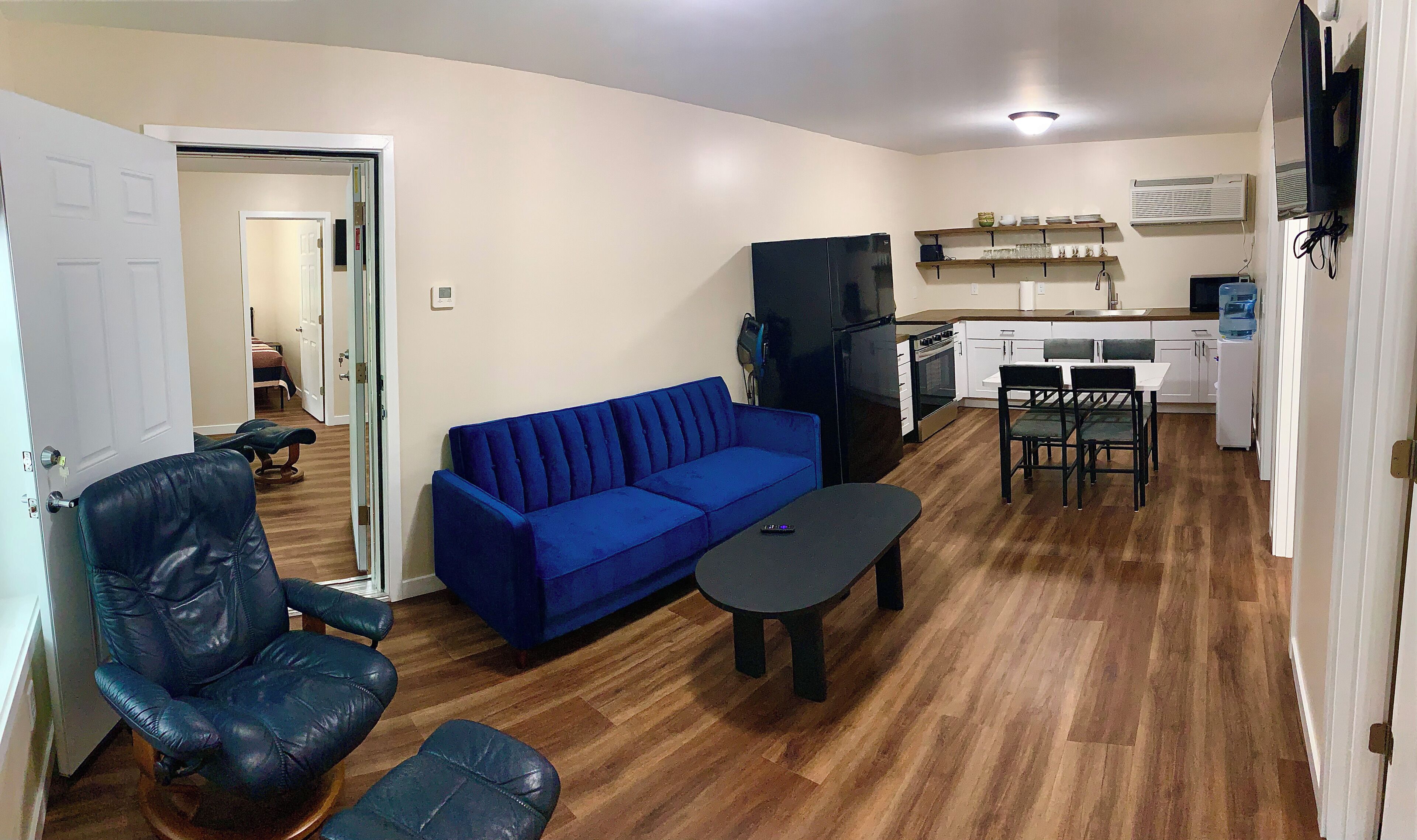 Un espacio habitable compacto con un sofá azul, un sillón de cuero negro, una pequeña mesa de comedor y una zona de kitchenette.